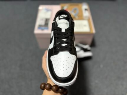 A hand holding a black and white Nike Dunk Low sneaker with a shoebox in the background.