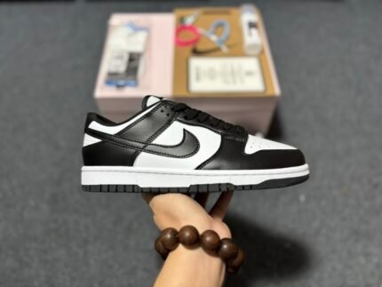 A hand holding a black and white Nike Dunk Low sneaker with a shoebox in the background.