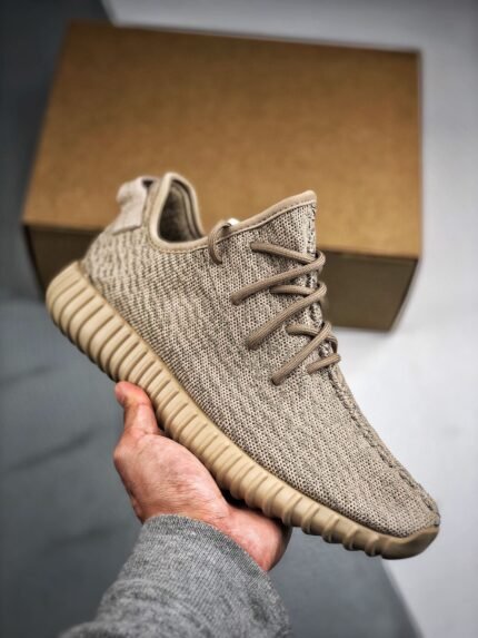 In the image, a hand is holding a beige sneaker with a brown shoebox visible in the background. The shoe features a Primeknit upper with a fine texture that is both breathable and contoured to the foot. The laces match the upper's color, contributing to a minimalist and elegant design. The outsole has a distinctive pattern and is colored to complement the upper, offering good cushioning and traction. The design of these shoes emphasizes comfort and style, making them suitable for casual everyday wear.