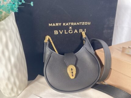 A dark blue handbag with a gold snake head decoration and chain shoulder strap, with a dark box and white vase in the background.