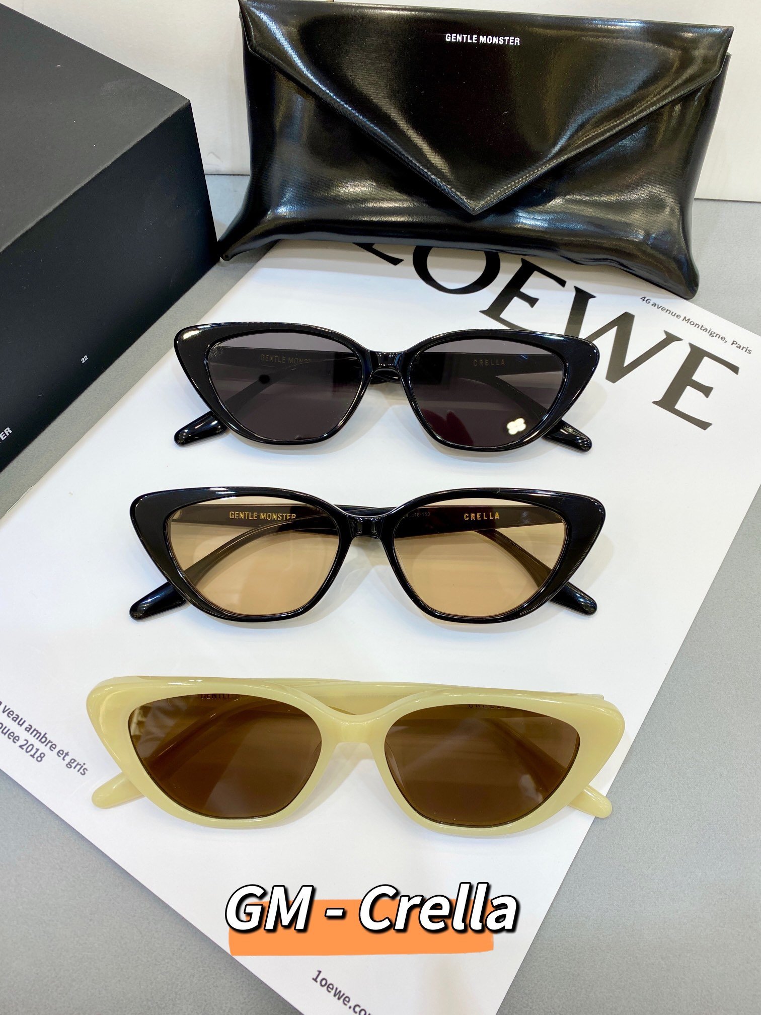 Gentle Monster Crella Sunglasses Collection The image shows a collection of three pairs of Gentle Monster Crella sunglasses displayed on a white surface. The sunglasses come in different colors: black, brown, and cream. Each pair has a unique design, and the brand name "Gentle Monster" is visible on the frames. In the background, there is a black leather pouch and a magazine with the "LOEWE" logo.
