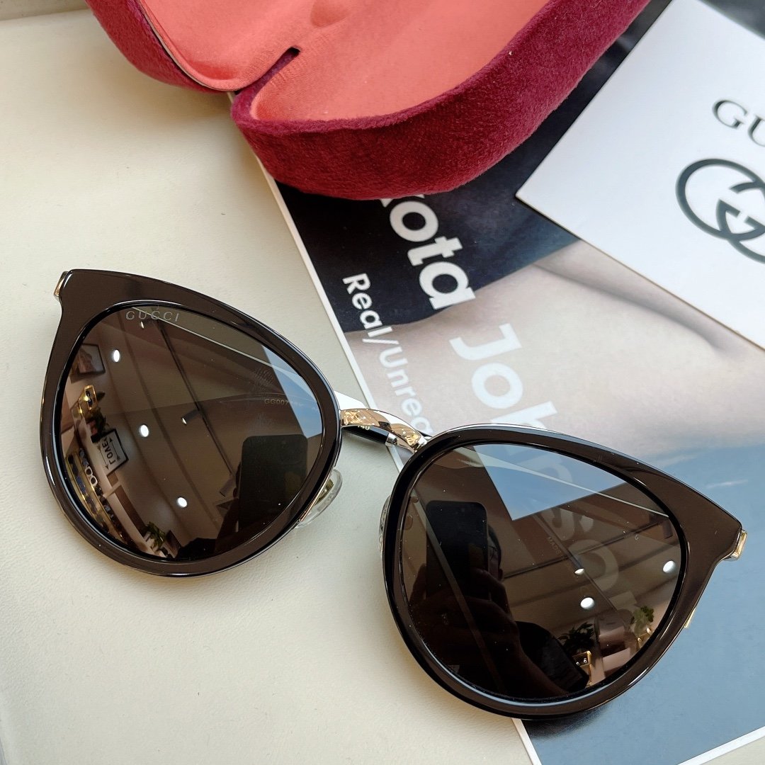 A pair of black Gucci sunglasses with the brand logo on the lenses, placed on a light-colored surface next to a red velvet pouch and a magazine featuring the Gucci logo.