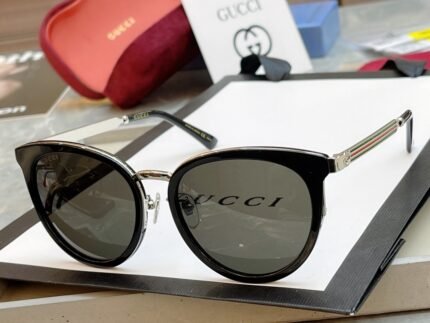 A pair of black Gucci sunglasses with the brand logo on the lenses, placed on a white surface. A red Gucci glasses case and a booklet with the Gucci logo are visible in the background.