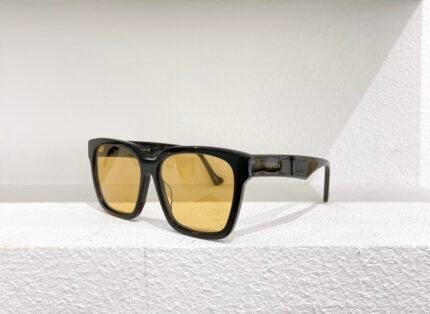 A pair of black Gucci sunglasses with yellow lenses, placed on a white surface against a white wall.