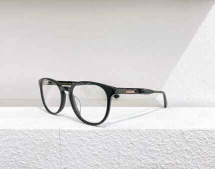 Black Gucci optical glasses with clear lenses, placed on a white surface against a white wall.
