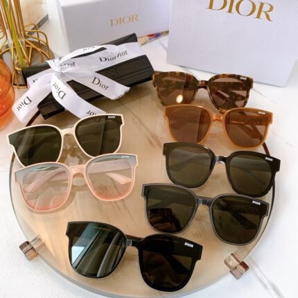 The image features a collection of Dior sunglasses displayed on a transparent tray. There are various styles and colors, including black, brown, and pink frames. The sunglasses are arranged neatly, and in the background, there are Dior branded boxes and ribbons, adding a luxurious feel to the presentation.