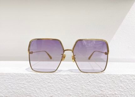 A pair of stylish sunglasses with a gold frame and gradient lenses, placed on a white textured surface against a white background.