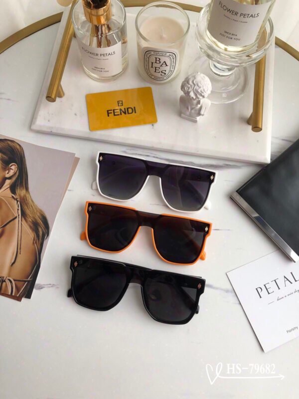 The image shows a collection of stylish sunglasses displayed on a marble surface. There are three pairs of sunglasses in different colors: black, white, and orange. In the background, there are decorative items such as a vase with flowers, a candle, and a Fendi branded card, creating an elegant and luxurious atmosphere.