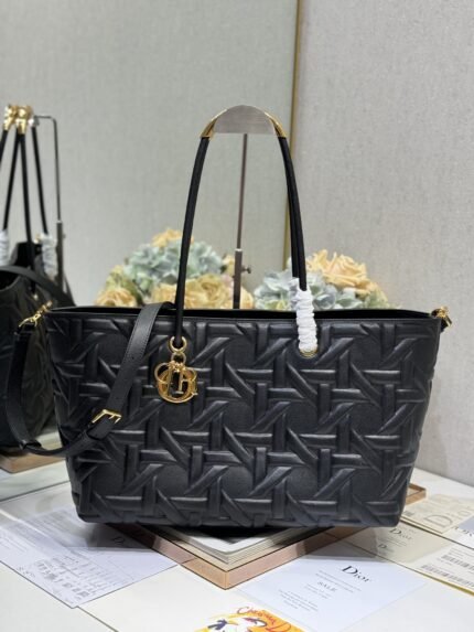 Black woven texture tote bag with gold charm, comes with a long shoulder strap, against a backdrop of brand books and packaging
