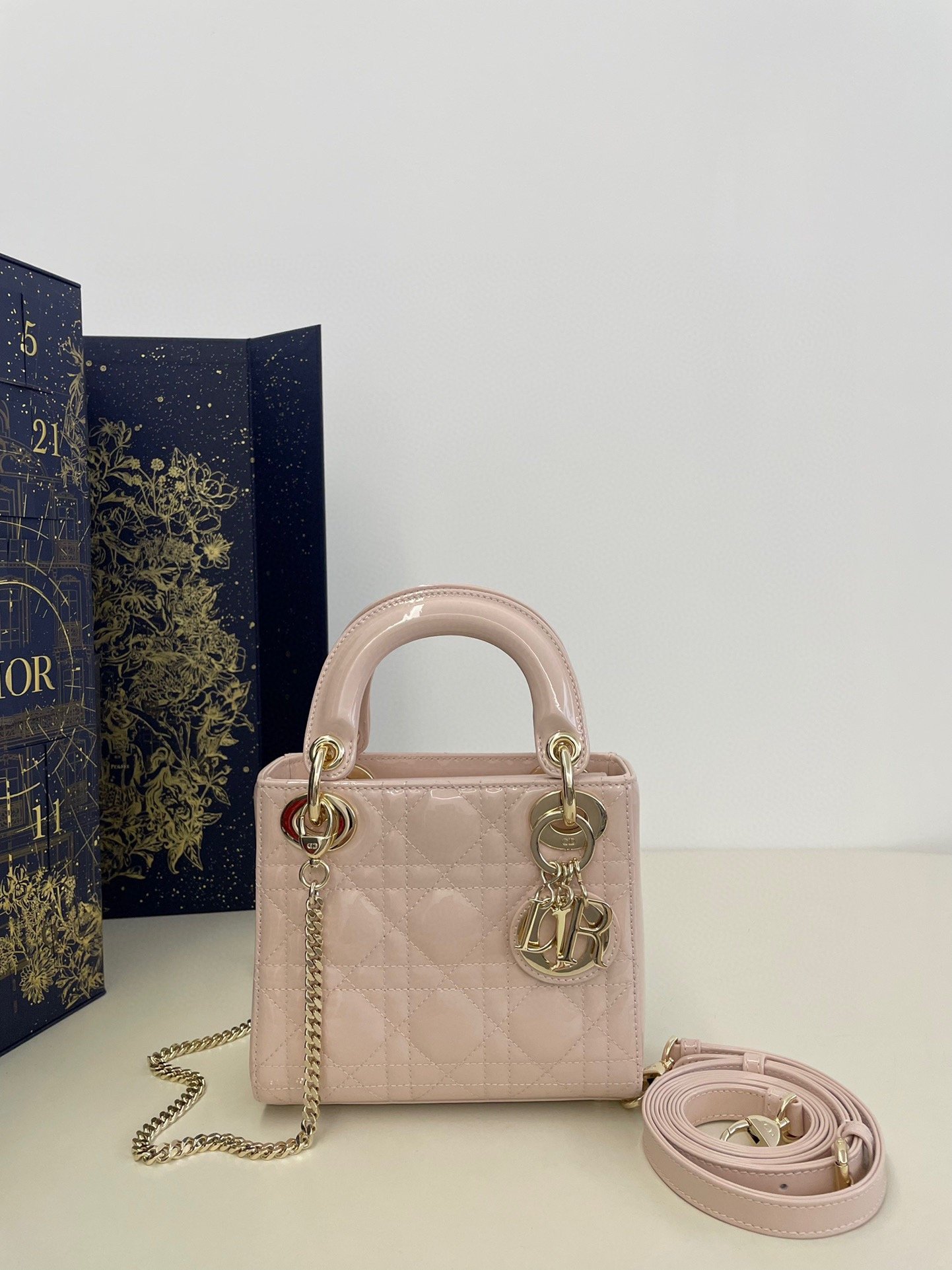 Pink patent leather quilted handbag with gold chain and charm, comes with a long shoulder strap, against a decorative box background