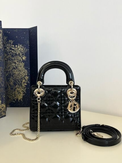 Black patent leather quilted handbag with gold chain and charm, comes with a long shoulder strap, against a decorative box background